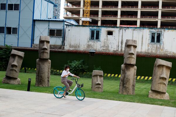 Jeune femme passant à vélo devant des reproductions de statues moaï, Pékin. - Sputnik Afrique