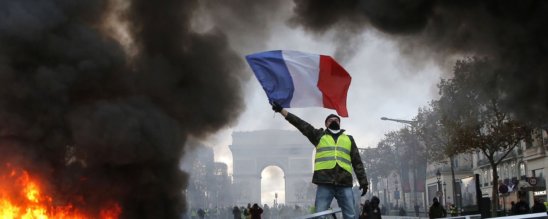 In this Nov. 24, 2018 file photo, a demonstrator waves the French flag on a burning barricade on the Champs-Elysees avenue with the Arc de Triomphe in background, during a demonstration against the rise of fuel taxes.  - Sputnik Africa, 1920, 01.05.2023
