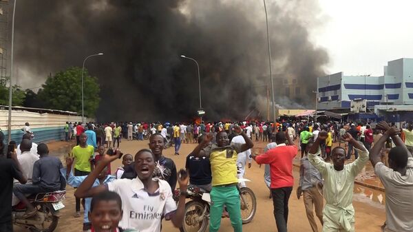 With the headquarters of the ruling party burning in the back, supporters of Niger's ruling junta demonstrate in Niamey, Niger - Sputnik Africa