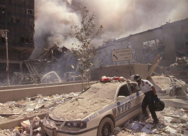 Rescue worker reaching into a New York Police car covered with debris while New York City fire fighters spray water on smoldering ruins in background, following September 11th terrorist attack. - Sputnik Africa