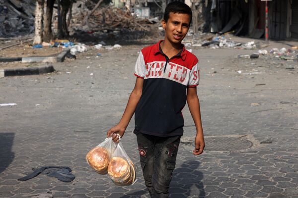 A young boy carries bags of bread as he walks in front of a building damaged by Israeli strikes in Gaza City on October 28, 2023. - Sputnik Africa
