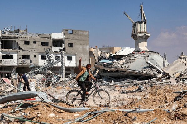 A man gestures as he rides a bicycle along a heavily damaged road past destroyed buildings in Khan Yunis on April 7, 2024 after Israel pulled its ground forces out of the southern Gaza Strip. - Sputnik Africa