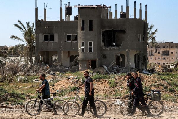 Men walk with bicycles along a heavily damaged road past a destroyed building in Khan Yunis on April 7, 2024 after Israel pulled its ground forces out of the southern Gaza Strip. - Sputnik Africa