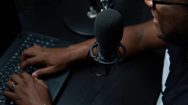 Close-up of an African podcaster blogger smiling during broadcast of his live audio podcast in studio. Male radio host with glasses, using a microphone and headphones, conducts a podcast or interview - Sputnik Africa