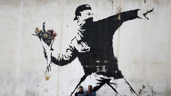 Two men are sitting in front of a famous graffiti of British street artist Banksy, painted on a wall of a gas station in the West Bank city of Bethlehem on December 16, 2015. - Sputnik Afrique