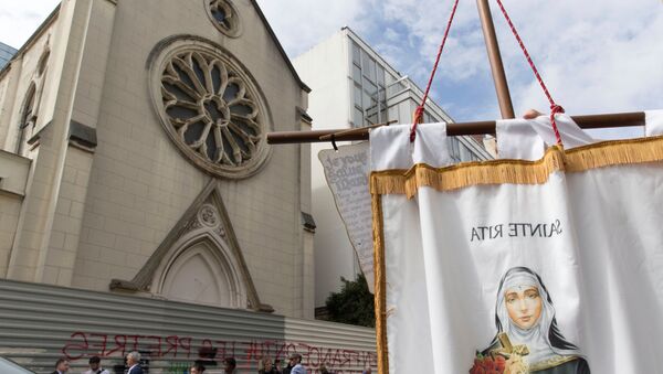 A person holds a banner picturing Sainte-Rita as other stand in front of barriers blocking the access to the sainte-Rita church in Paris with an inscription on it which translates as In France, priests are killed and churchs are demolished, on August 3, 2016 - Sputnik Afrique