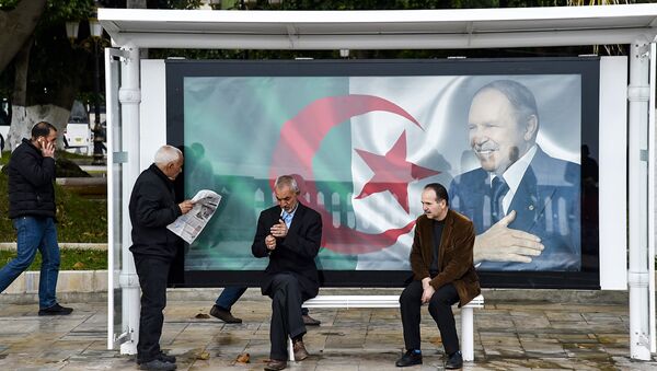 An Algerian reads a newspaper at a bus station next to a banner showing the Algerian flag with a portrait of President Abdelaziz Bouteflika, in the capital Algiers on February 11, 2019, as the country prepares for the upcoming presidential election scheduled for April 18. - Sputnik Afrique