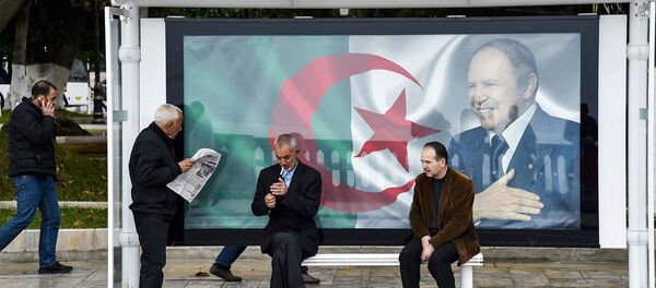 An Algerian reads a newspaper at a bus station next to a banner showing the Algerian flag with a portrait of President Abdelaziz Bouteflika, in the capital Algiers on February 11, 2019, as the country prepares for the upcoming presidential election scheduled for April 18. - Sputnik Afrique